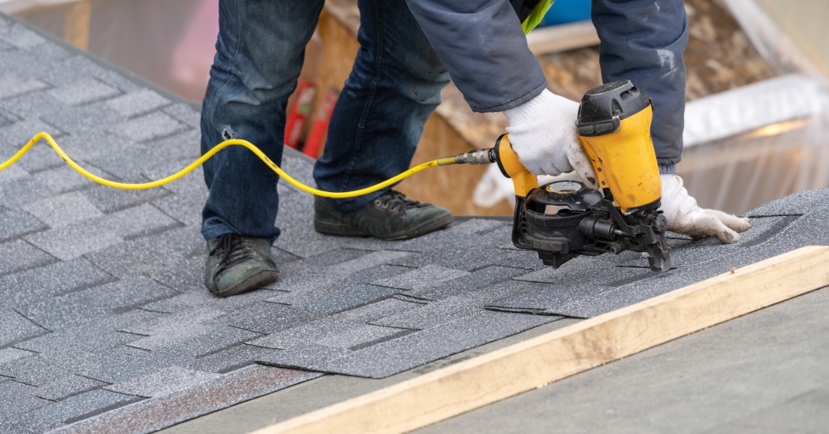 A close-up of a worker using a yellow pneumatic nail gun to install a roof tile onto a residential roof.