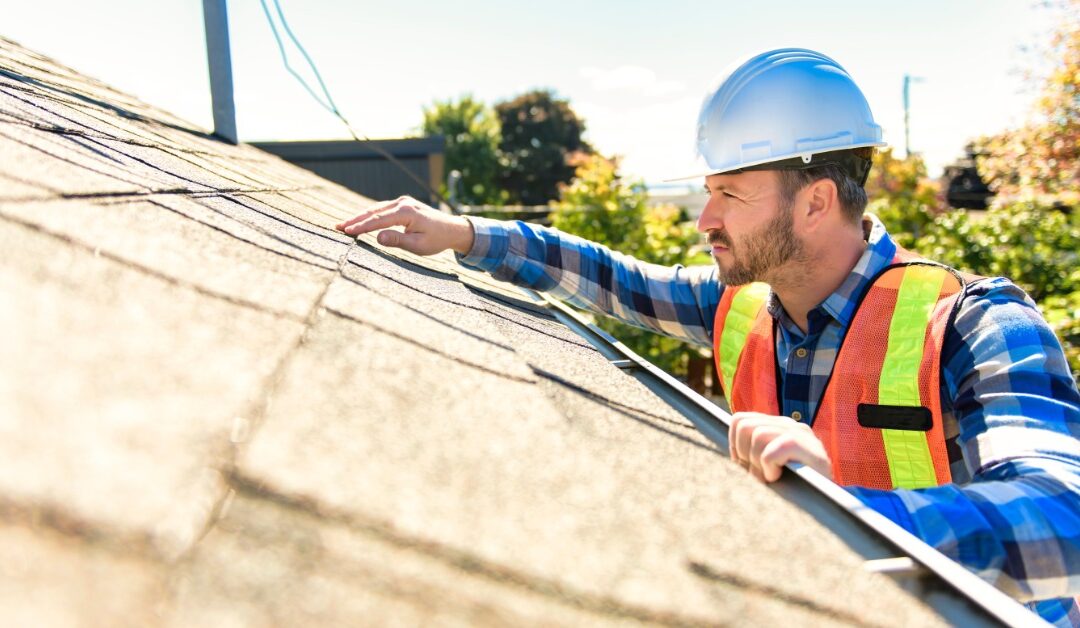 A close-up of a man in a flannel shirt with a high-visibility vest and white hard hat inspecting a home's roof.