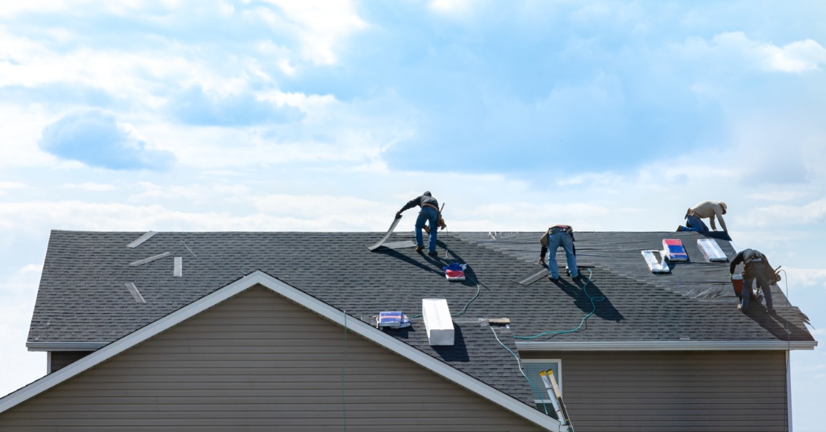 A distant view of four workers standing on a residential roof to install shingles with a cloudy sky behind them.