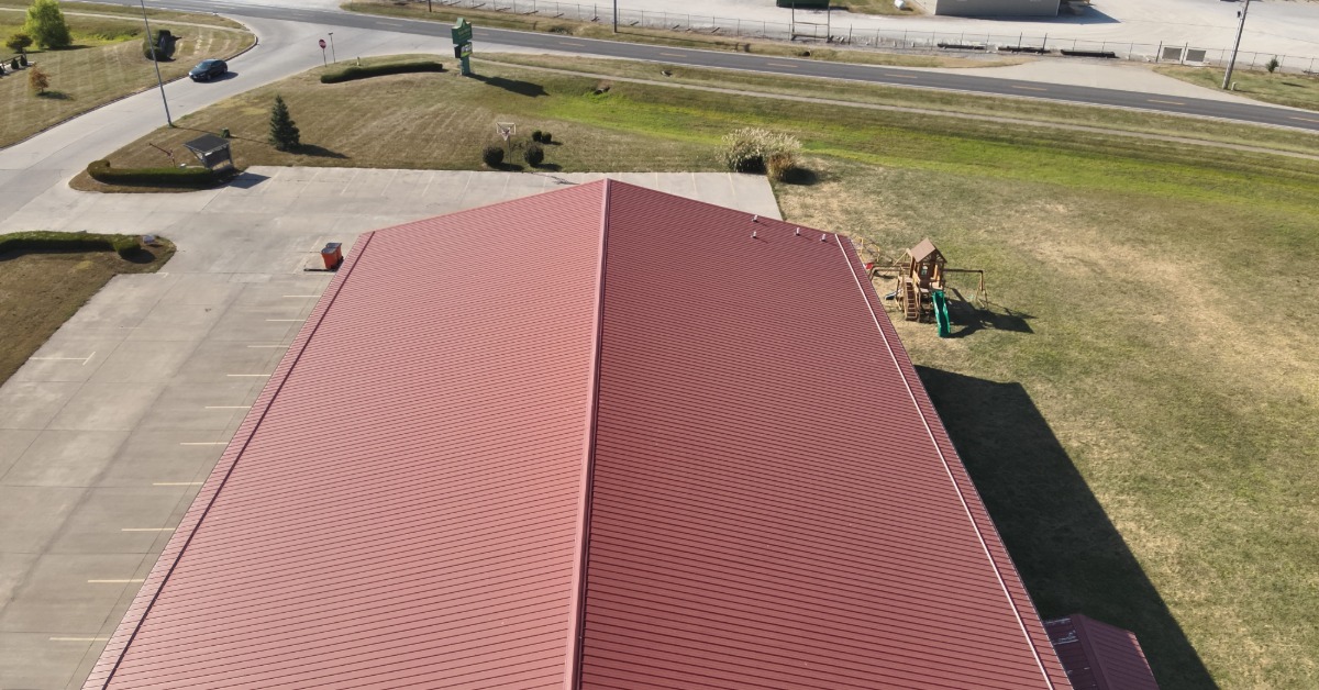 An overhead view of the red roof of a long, rectangular commercial building with a parking lot on a sunny day.