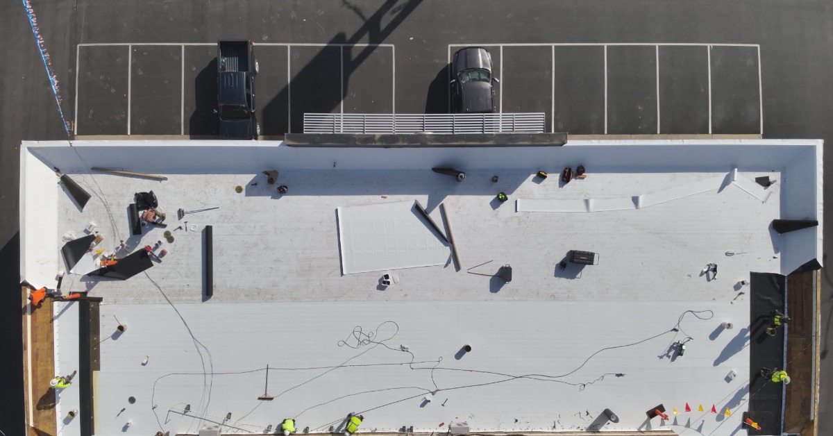 An overhead view of a commercial building with a black parking lot and multiple contractors working on the roof.