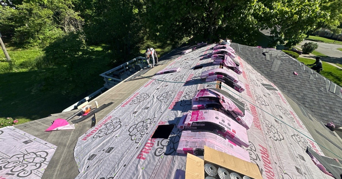 A roof covered with pink underlayment and rows of shingle bundles as a worker kneels near the roof edge.