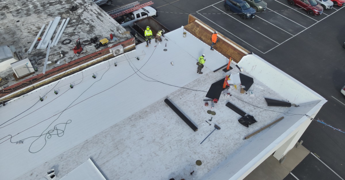 An aerial view of a large, white commercial building roof with workers on it and a large parking lot in the background.