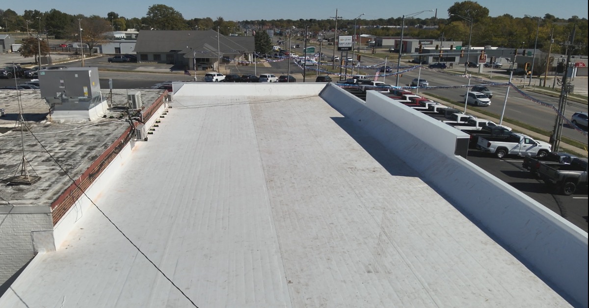 The roof of a commercial building. The white roof is mostly flat with a slight middle incline next to another building.