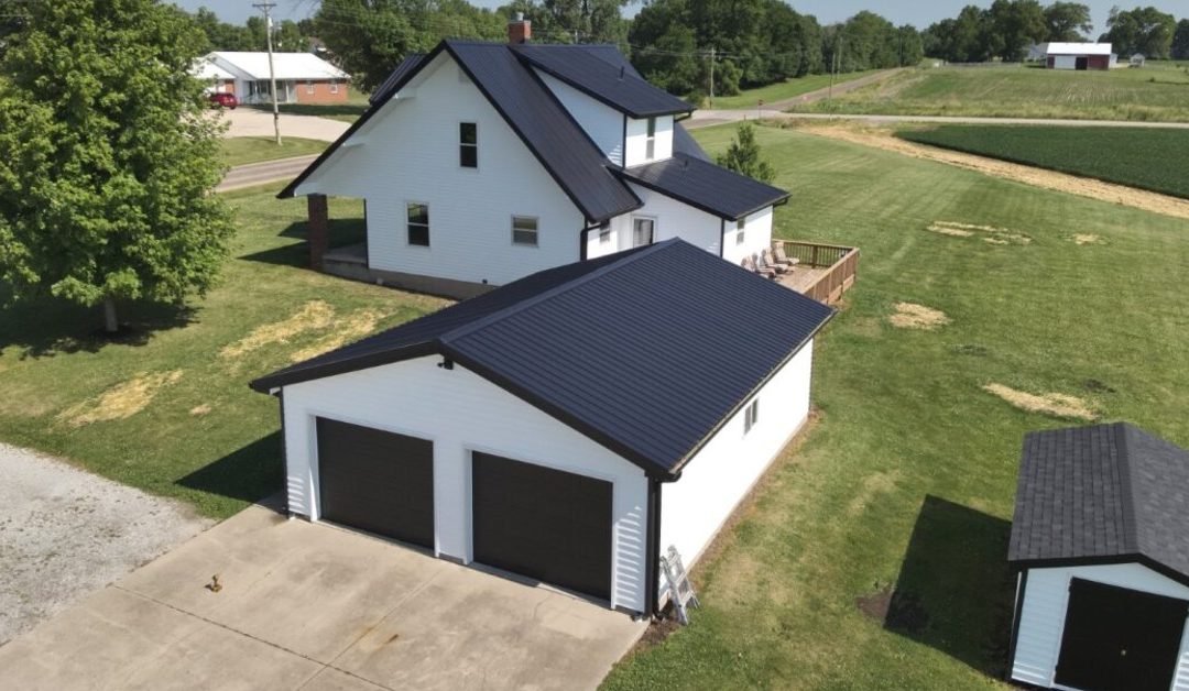An aerial view of a property with a home and a matching two-stall garage with white siding and a metal roof.
