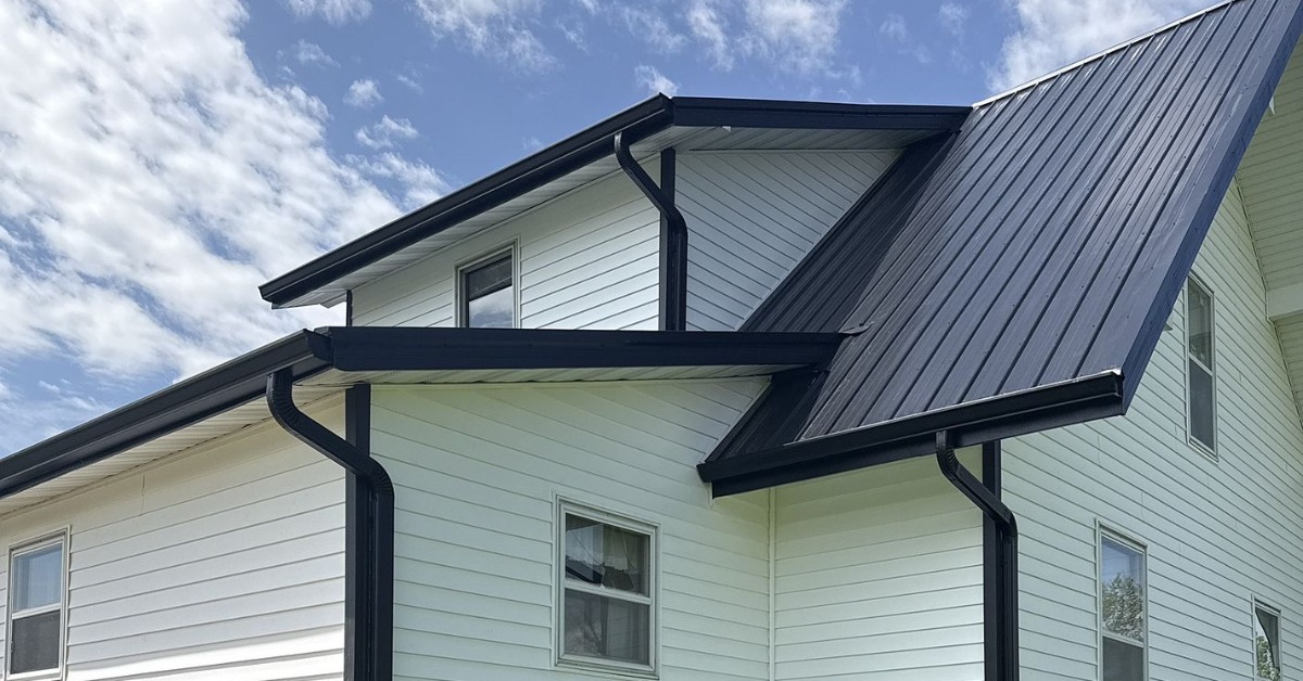 A ground-level view from the backyard of a home with a deck, white siding, and a metal roof during the day.