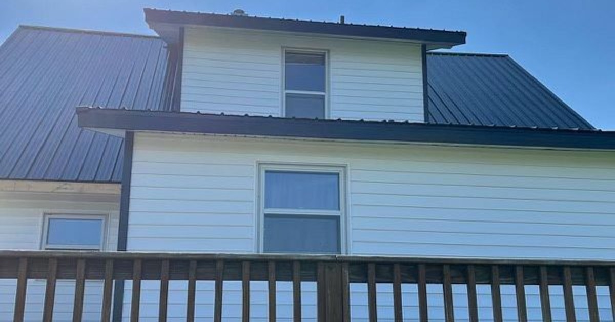 A view from the backyard looking up at a two-story home with a wooden deck, white siding, and a metal roof.