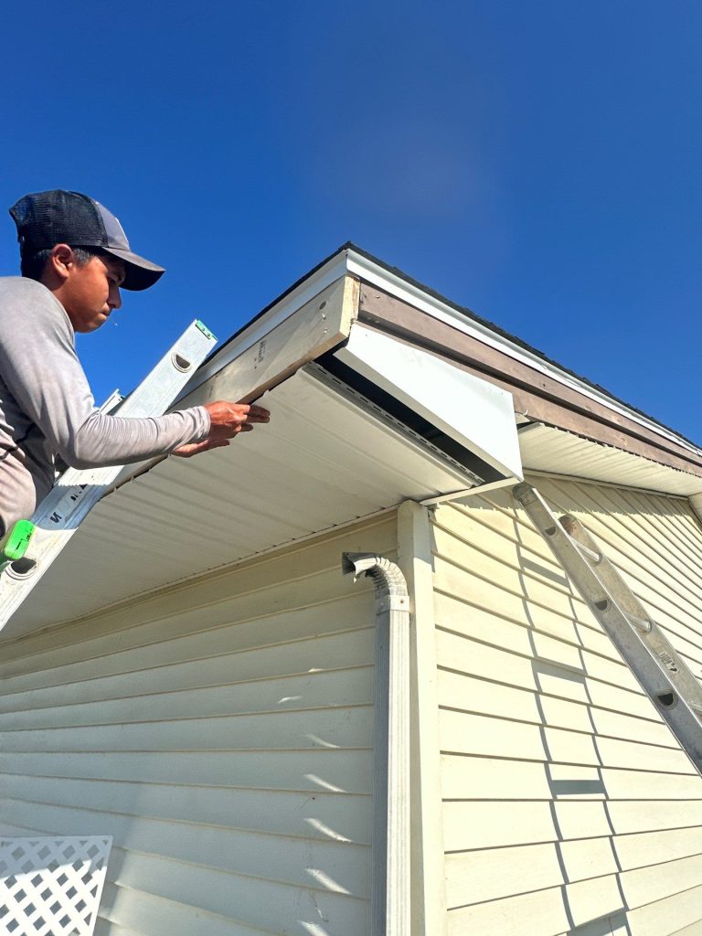 A close-up of a man on a ladder installing white vinyl material underneath the overhang of a home's roof.
